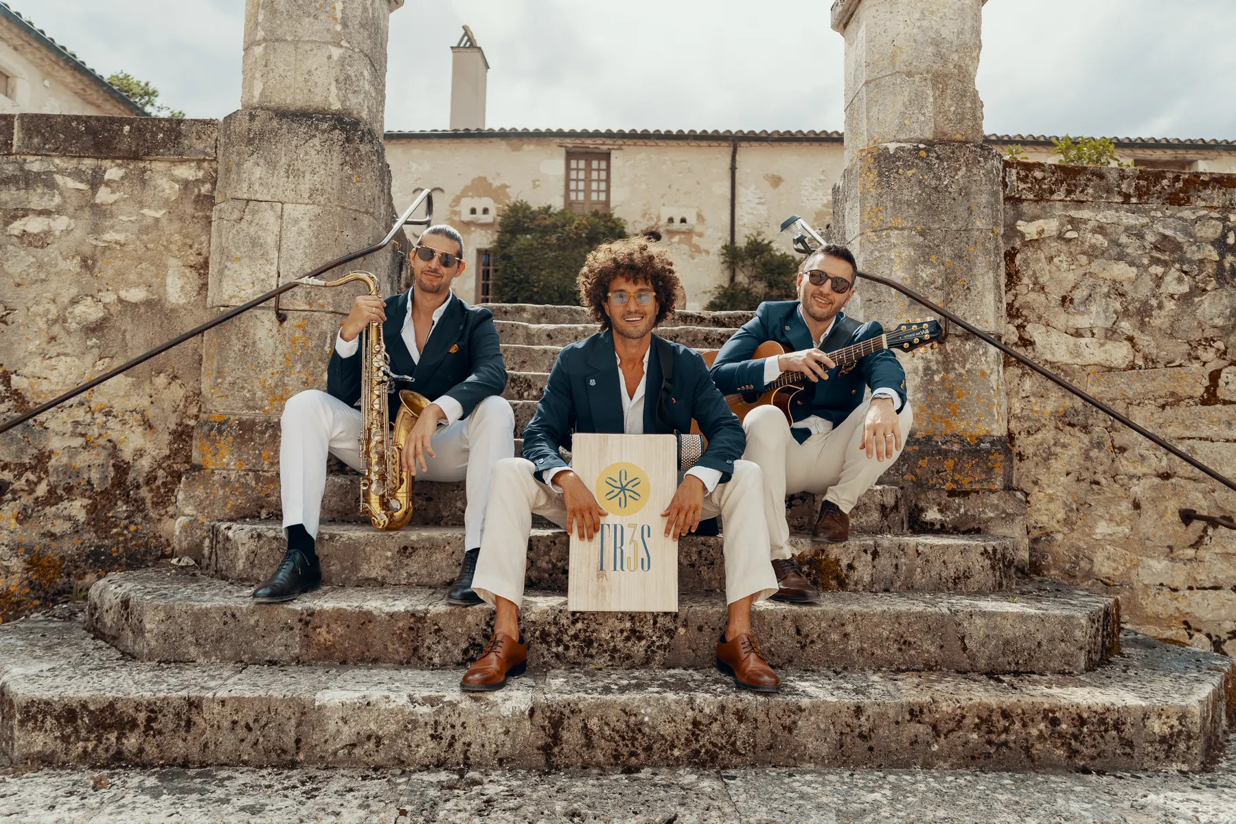 The three members of TR3S sitting on old stone steps looking at the camera, the saxophone, the guitar and the TR3S cajon facing us