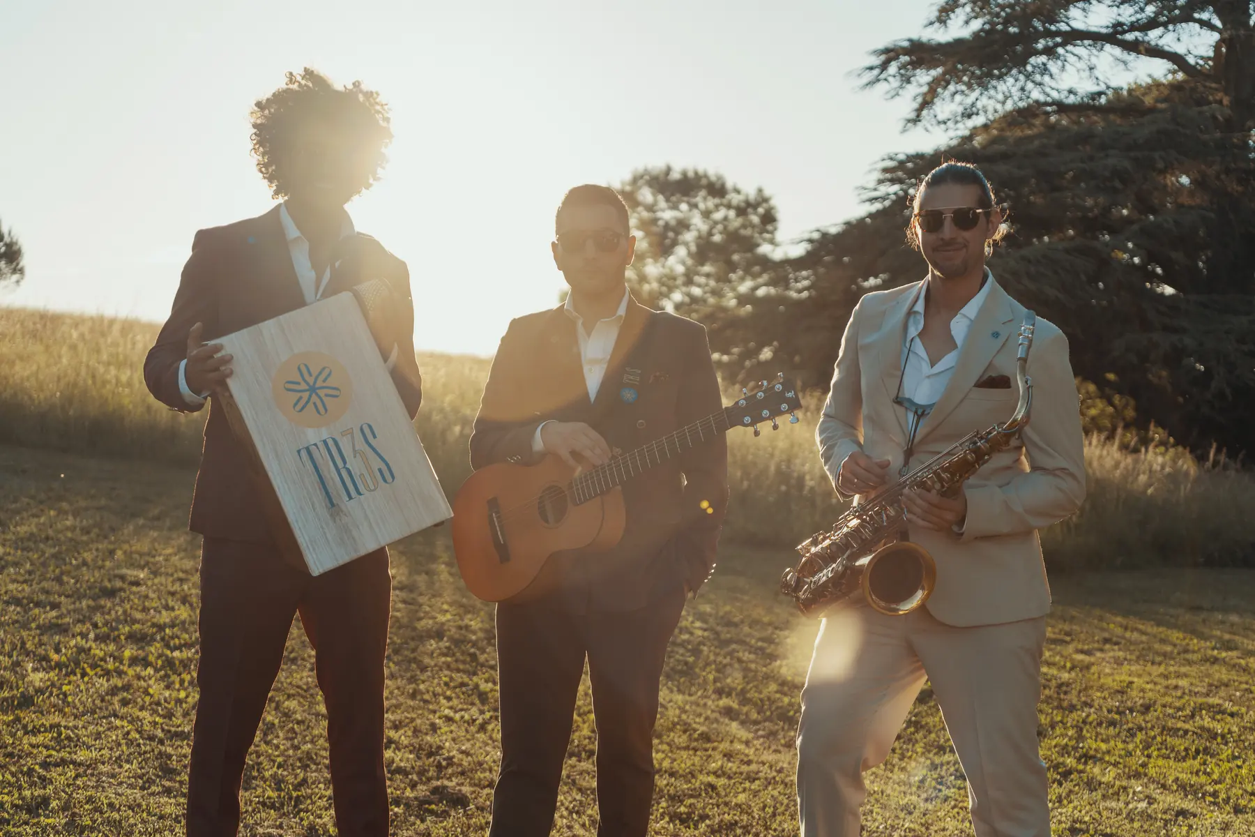 The three members of TR3S standing side by side in a golden field, the cajon logo facing the camera