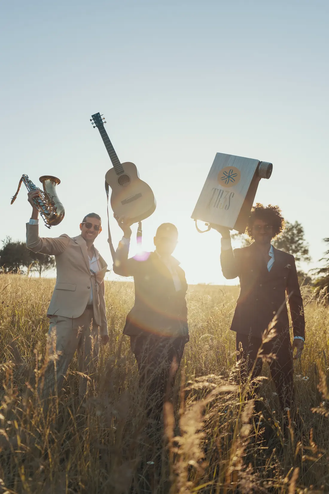 The trio raising a saxophone, a guitar and a cajon to the sky in tall grass at sunset