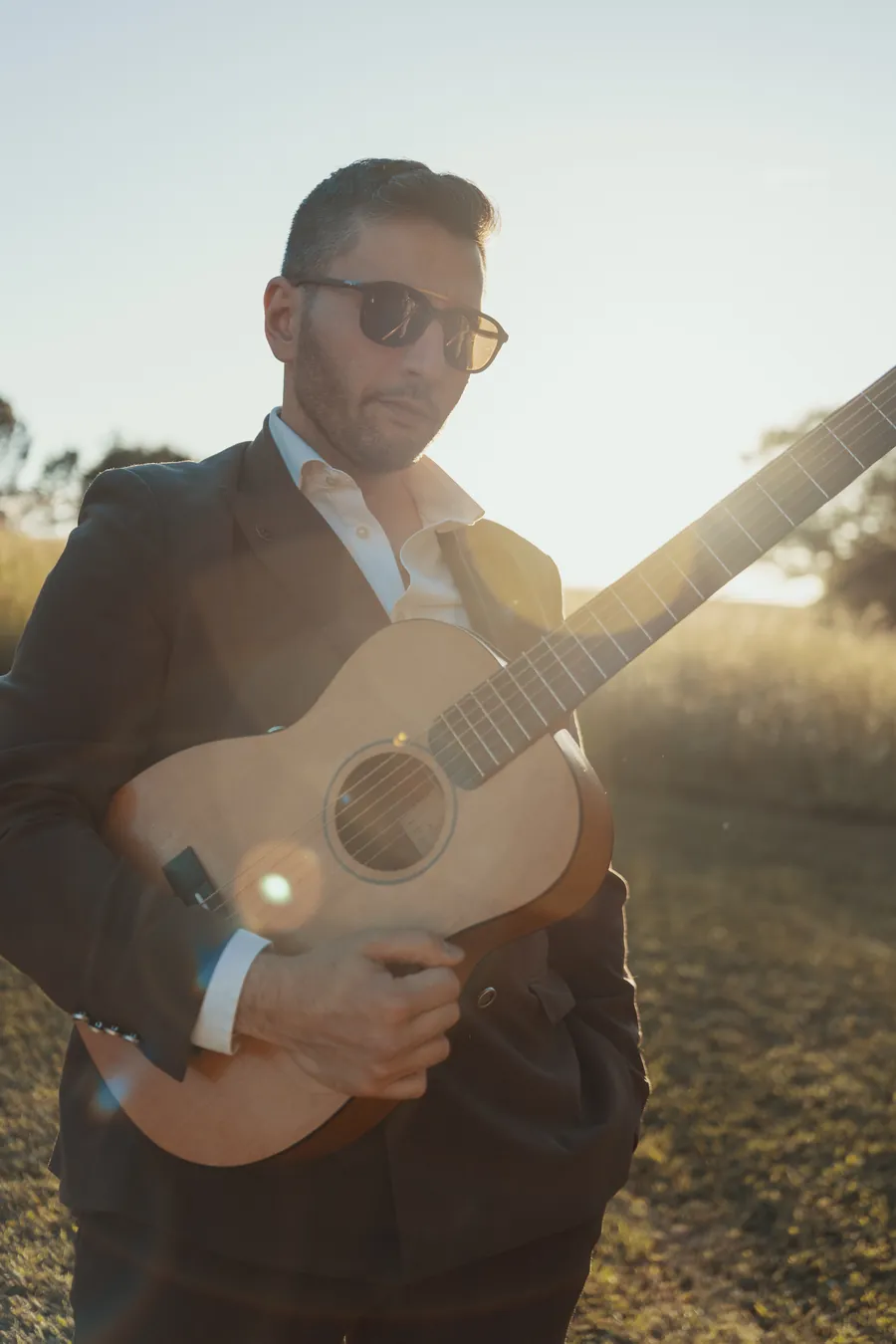 Frank, the TR3S guitarist, in sunglasses and a dark suit holding his acoustic guitar against the low sun