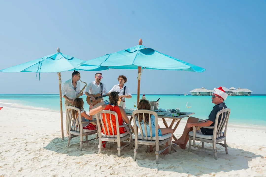Three TR3S musicians with a saxophone and guitar performing under turquoise parasols for a table of guests on a white sand beach in the Maldives