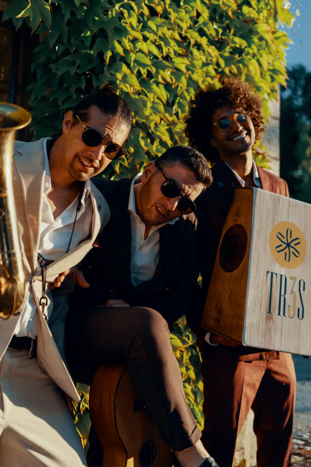 The three members of TR3S in sunglasses leaning together against an ivy wall, with a saxophone, an acoustic guitar and a cajon carrying the TR3S logo